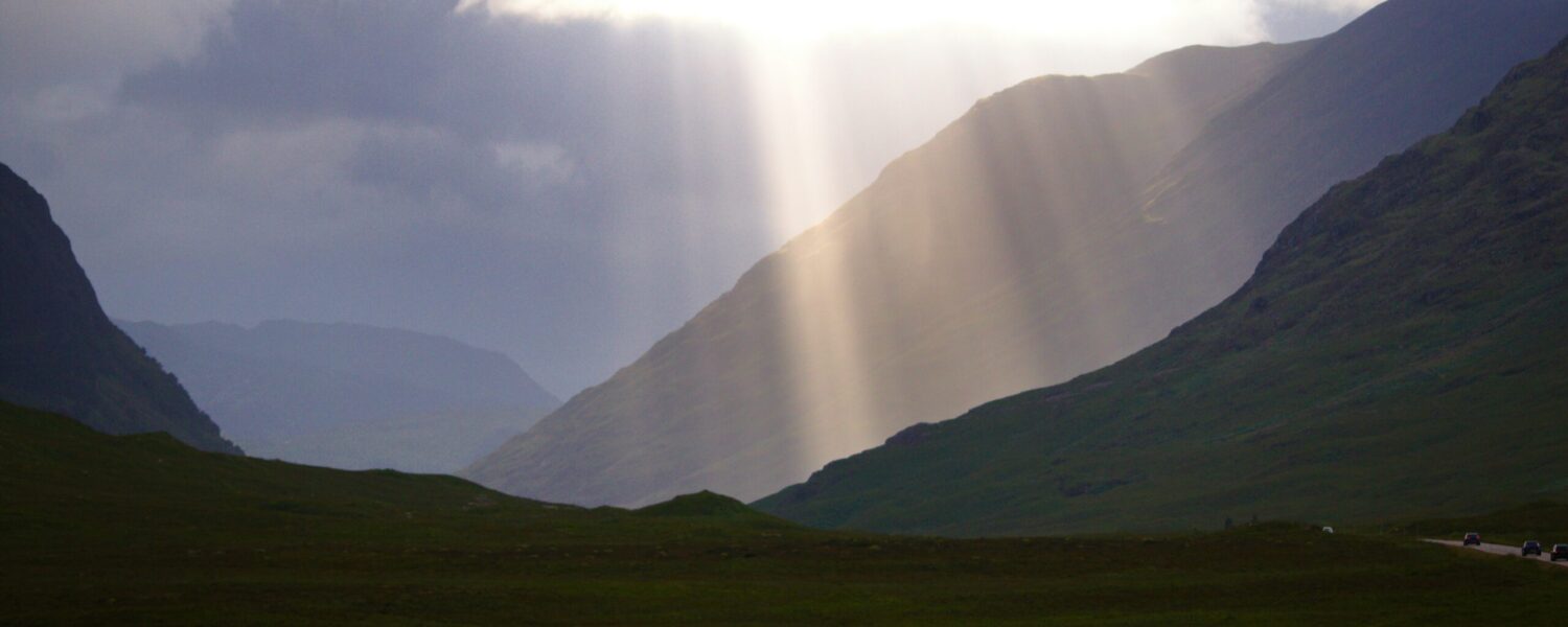 Sonnenlicht, das durch dunkle Wolken über einem grünen Tal und Berghängen bricht